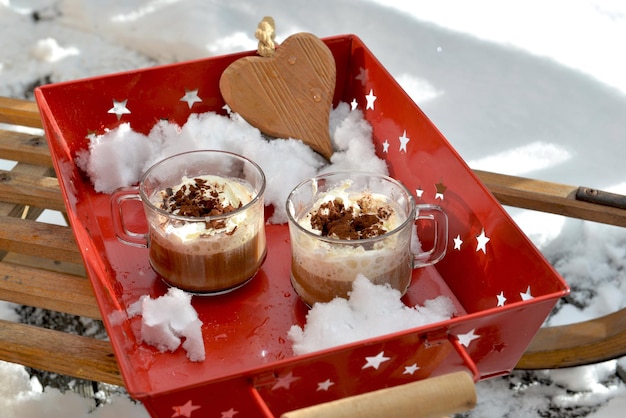 Duas xícaras de capuccino e coração de madeira colocadas em uma bandeja vermelha em um trenó de madeira na neve