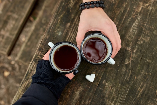 Duas xícaras de chá na natureza nas mãos de um casal em luvas pretas na superfície de madeira