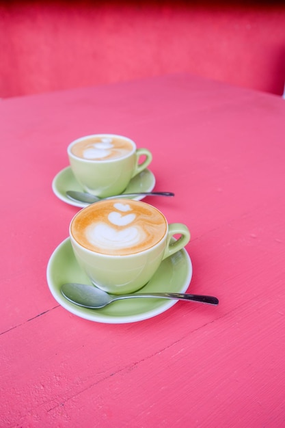 Duas xícaras verdes de cappuccino com latte art em fundo rosa pastel