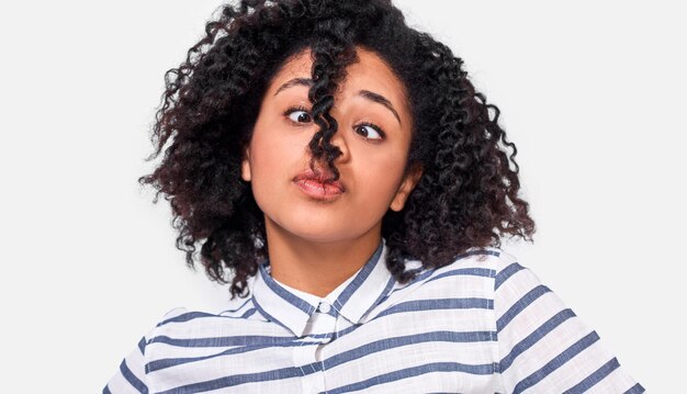 Dudosa joven afroamericana mirando a un lado con cara curiosa vestida con una camisa a rayas casual posando sobre una pared blanca La estudiante tiene una expresión confusa debido al peinado