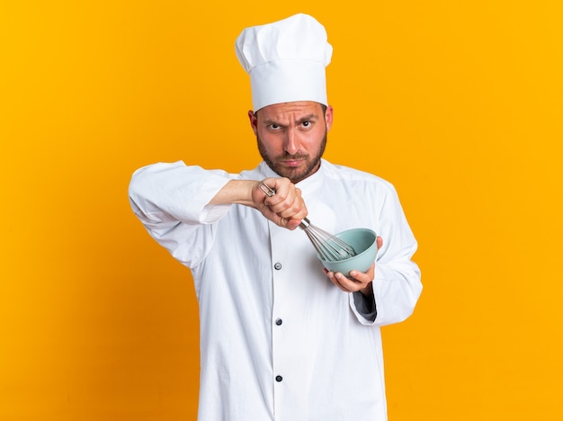 Dudoso joven cocinero de sexo masculino caucásico en uniforme de chef y gorra mirando a la cámara batiendo el huevo en un tazón con batidor aislado en la pared naranja