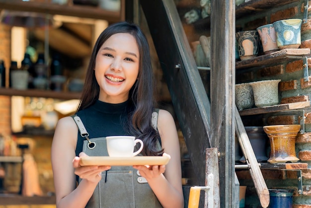 La dueña del café sonriendo con felicidad positiva lista para abrir para el cliente mujer asiática con delantal bandeja de regalo de mano y taza de café cálida bienvenida segura de pie en la entrada de la cafetería