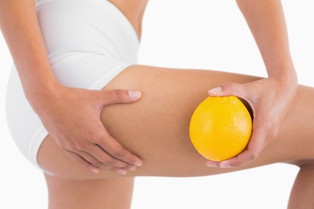 Dünne Frau, die Cellulite-Haut auf Schenkel zusammendrückt, wie sie Orange hält