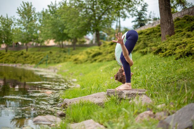 Dünnes Brunettemädchen spielt Sport und führt die schönen und hoch entwickelten Yogahaltungen in einem Sommerpark durch.