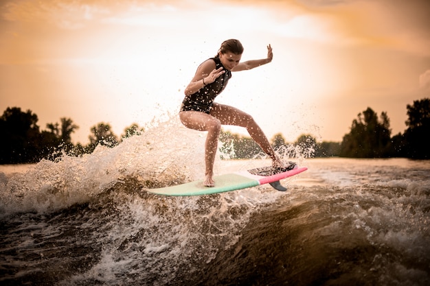 Dünnes Mädchen, das auf das Wakeboard auf dem Fluss auf der Welle im Sonnenuntergang springt