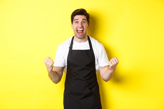 Dueño de la cafetería emocionado en delantal negro celebrando hacer puño y gritando de alegría lograr g ...