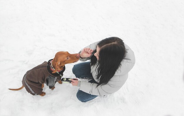 El dueño y un lindo perro marrón en la ropa se sientan en la nieve y juegan.