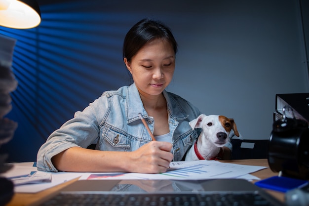 Dueño de la mujer asiática que trabaja horas extras con su perro en casa. Área de trabajo interesante de Jack Russell Terrier. Informe del documento de verificación. Trabajar desde casa.