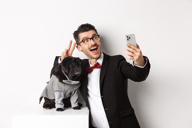 Dueño de perro alegre en traje celebrando el año nuevo con perro, tomando selfie en smartphone cerca de lindo pug negro en traje, fondo blanco.