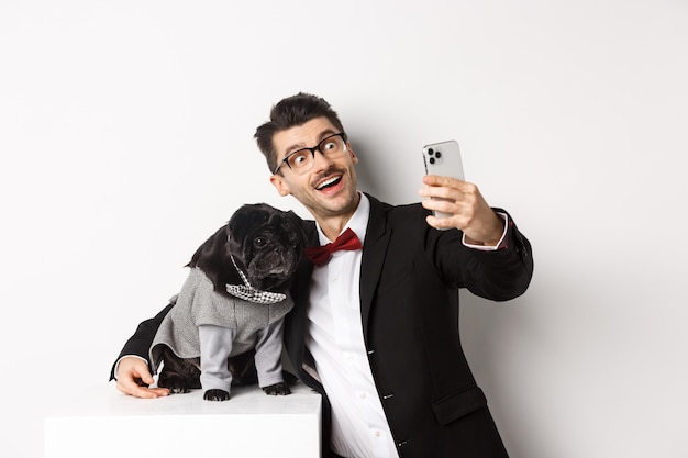 Dueño de perro alegre en traje celebrando la Navidad con perro, tomando selfie en smartphone y abrazando lindo pug negro en traje, fondo blanco.