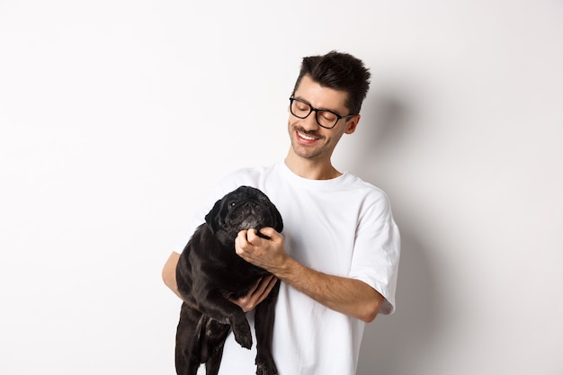 Dueño de perro feliz rascarse la cara de pug negro lindo y sonriente, acariciar a su animal, de pie sobre fondo blanco.