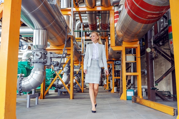 Dueño de la planta de energía mujer rubia sonriente caminando y comprobando la maquinaria.