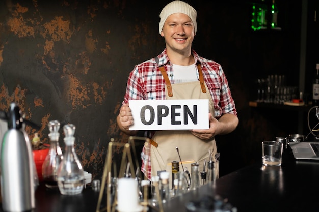 El dueño sonriente del bar se para detrás de la barra y sostiene el cartel abierto Invita a todos a entrar Abierto