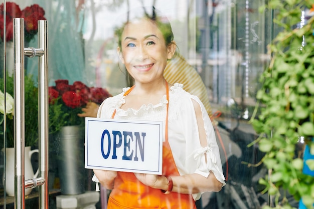 Dueño de la tienda de flores senior femenino sonriente en delantal naranja girando el letrero abierto en la puerta de vidrio