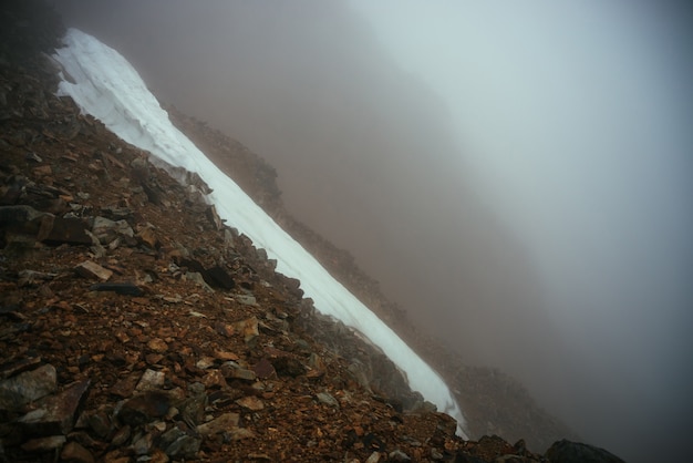 Düstere minimalistische neblige Berglandschaft mit Schneegesims über Abgrund in der Wolke. Über Schneegesims am Abgrund im dichten Nebel. Gefährlicher Gebirgspass über dem Abgrund. Gefahr Felsen im dichten Nebel.