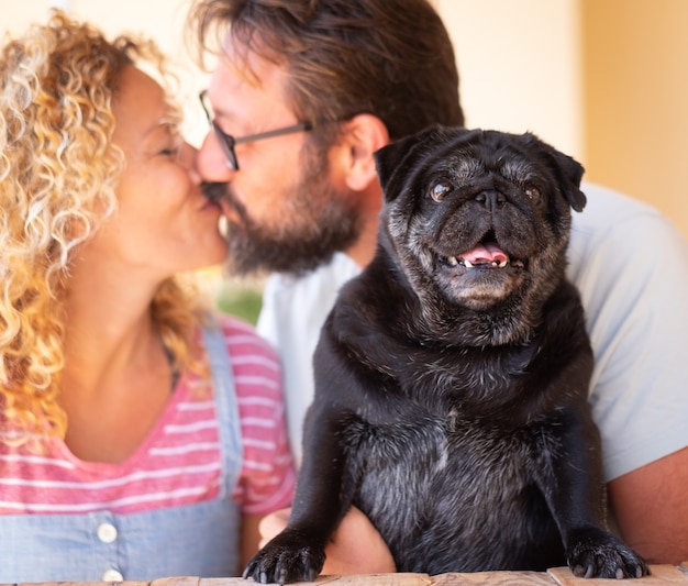 Un dulce beso entre esposa y esposo abrazando a su viejo perro pug negro al aire libre en la terraza