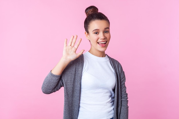 Dulce hola de una chica encantadora. Retrato de una amigable adolescente morena con peinado de moño con ropa informal saludando con la mano, saludando, saludando y sonriendo a la cámara. tiro del estudio aislado, fondo rosa