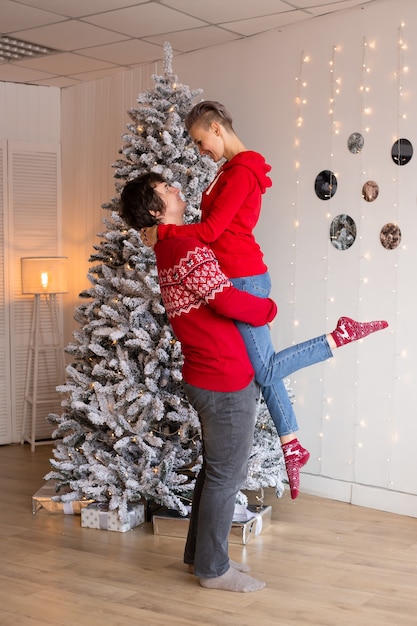 Dulce joven pareja de enamorados besándose en la víspera de Navidad cerca del árbol con regalos en casa en diciembre.