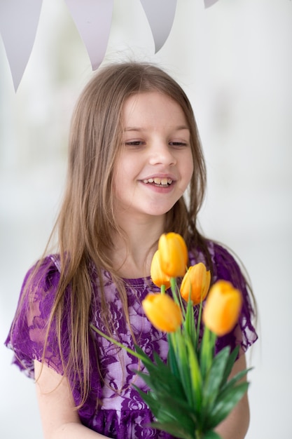 Dulce niña de cabello oscuro en vestido morado en estudio con tulipanes amarillos