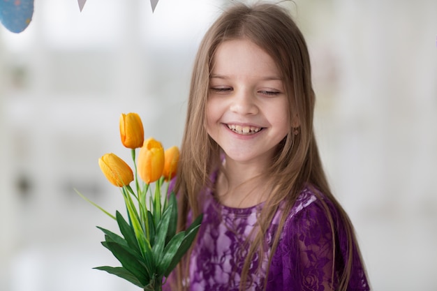 Dulce niña de cabello oscuro en vestido morado en estudio con tulipanes amarillos