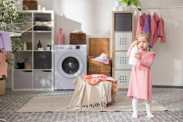 Una dulce niña juega en el baño de la lavandería junto a la cesta de la ropa con ropa