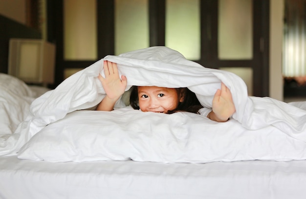 Dulce niña mirando a cámara y sonriendo mientras está acostado en la cama debajo de la manta