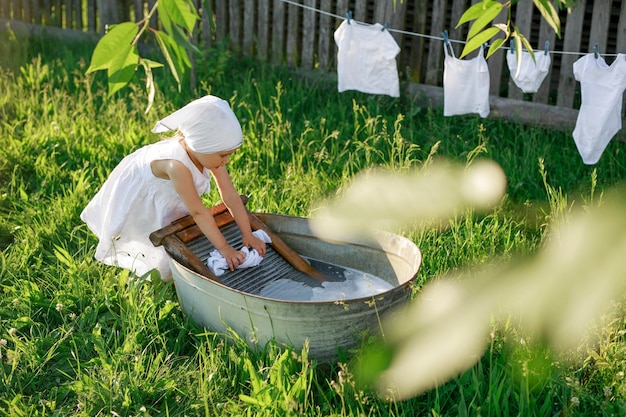 Dulce niña en un pañuelo y un vestido de lavado de lino blanco como la nieve a mano en una vieja tabla de lavar