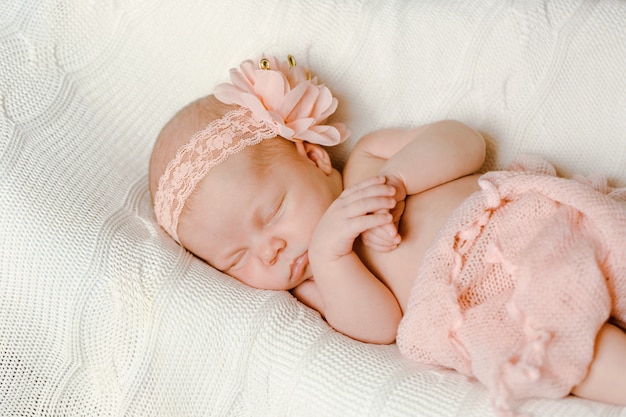 Una dulce niña recién nacida, envuelta en una suave manta rosa con una venda rosa, duerme sobre una manta blanca de punto.