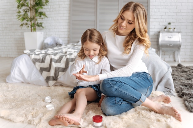 Dulce niña sentada en los abrazos de su madre y aplicando un poco de crema en su mano.