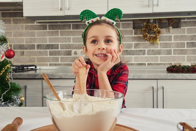 Dulce niño, adorable niña encantadora sonríe, mezclando ingredientes con piso en el tazón de vidrio. Pequeño chef preparando masa para galletas y pan de Navidad