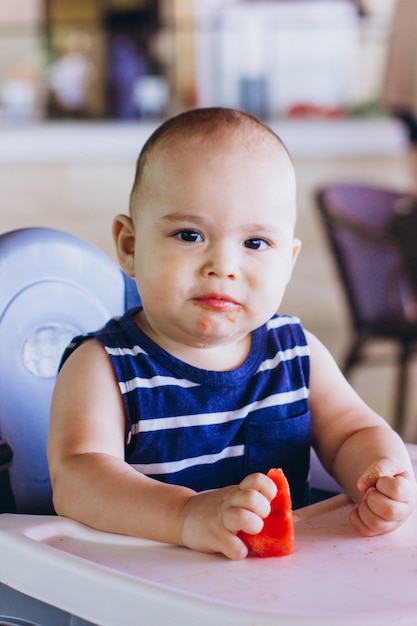 Dulce niño comiendo sandía roja sentado en la silla de bebé en el restaurante. Vacaciones de hotel con niño