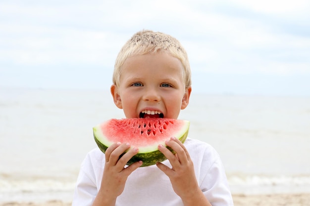 Dulce niño rubio en la costa sosteniendo una jugosa rebanada de sandía