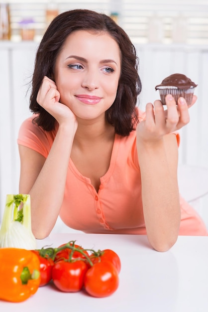 Dulce tentación. Mujer joven pensativa sosteniendo muffin de chocolate y mirándolo mientras las verduras frescas ponen cerca de ella en la mesa