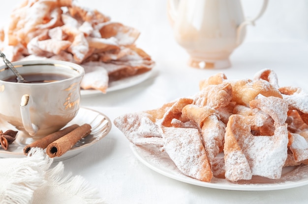 Dulces de carnaval de Chenchi en un plato blanco. Espolvoreado con azúcar en polvo. Cerca hay una taza de té decorada con canela. Fondo blanco.