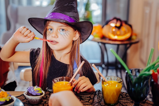 Dulces y jugos. Linda chica hermosa con traje de Halloween de mago disfrutando de dulces temáticos y jugo de naranja