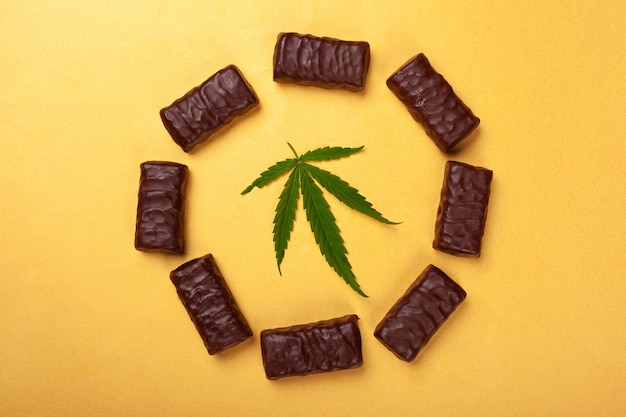Dulces con marihuana, círculo de chocolates con una hoja de cannabis medicinal en una vista superior de fondo amarillo.