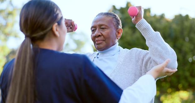 Dumbbell fitness e uma mulher negra sênior com uma enfermeira ao ar livre em um jardim juntos para fisioterapia Exercício de saúde ou bem-estar com um paciente idoso e voluntário no quintal para se exercitar