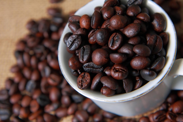 Dunkel geröstete Kaffeebohnen in weißer Tasse mit verschwommenen Kaffeebohnen und Gunny-Sack