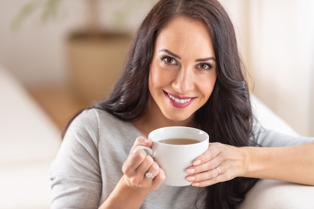 Dunkelhaarige Frau, die eine schöne Tasse Tee oder Kaffee genießt, während sie auf einer Couch sitzt.