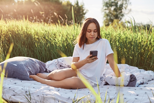 Dunkelhaarige schöne Frau mit weißem T-Shirt und kurzer Pose in der Mitte des Feldes mit Smartphone in der Hand, mit Handy, E-Mail checken, während sie sich in der Natur ausruht.
