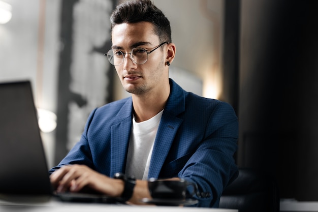 Dunkelhaariger junger Architekt mit Brille in blau karierter Jacke arbeitet am Laptop im Büro.