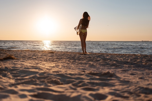 Dunkelhaariges Mädchen im Badeanzug steht am Sandstrand am Meer bei Sonnenuntergang und hält ein Surfbrett.