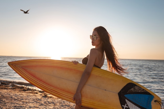 Dunkelhaariges Mädchen in Badeanzug und Sonnenbrille geht bei Sonnenuntergang am Sandstrand und hält ein Surfbrett.