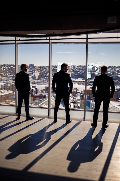 Dunkle männliche Silhouetten vor dem Hintergrund eines Panoramafensters. Männliche Geschäftsleute mit Blick aus dem großen Fenster eines Wolkenkratzers mit Blick auf die Metropole looking
