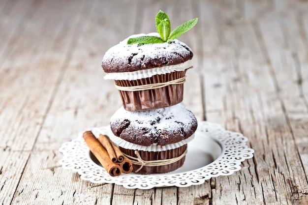 Foto dunkle schokoladenmuffins mit zuckerpulver, zimtstangen und tadellosem blatt auf weißer platte