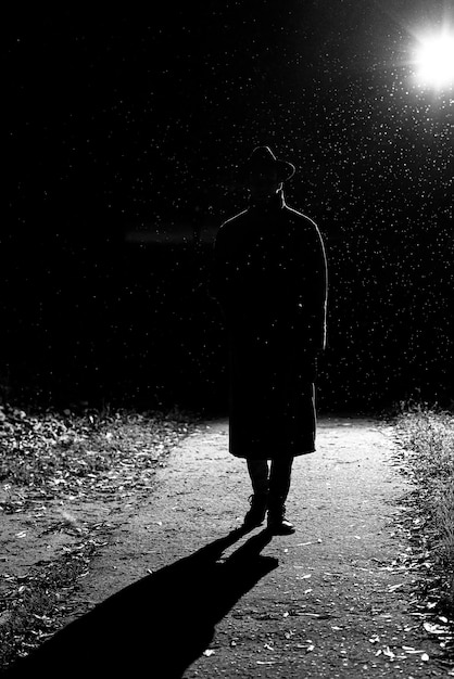 Dunkle Silhouette eines Mannes in einem Regenmantel und in einem Hut auf der Straße