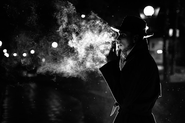 Dunkle Silhouette eines Mannes mit Hut Rauchen einer Zigarette im Regen auf einer Nachtstraße im Stil von Noir