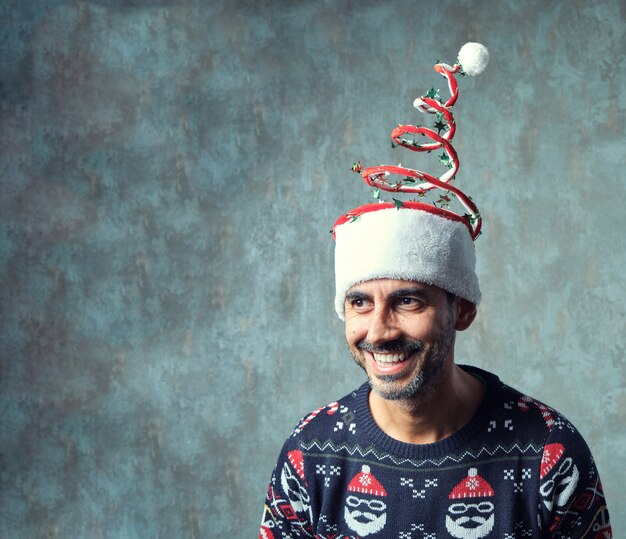 Dunkler Kerl mit kurzem grauem Bart, der in blauem Weihnachtspullover und lustigem Weihnachtshut lächelt