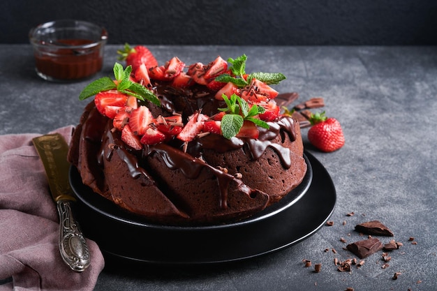 Dunkler Schokoladen-Bundt-Kuchen mit Ganache-Zuckerguss und Erdbeere auf dunklem Stein- oder Betontischhintergrund Festlicher Kuchen Selektiver Fokus