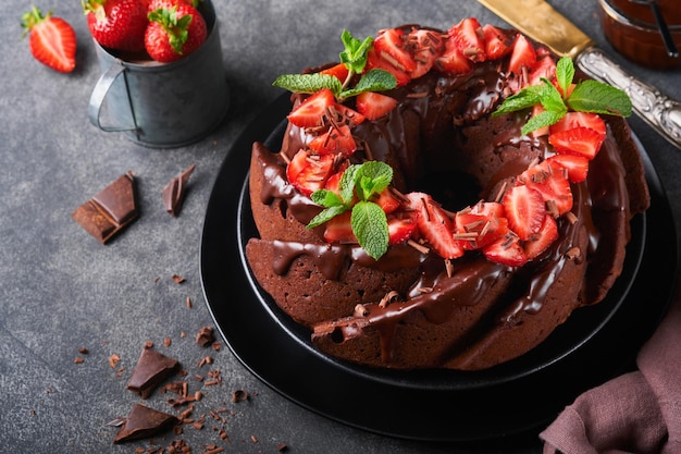 Dunkler Schokoladen-Bundt-Kuchen mit Ganache-Zuckerguss und Erdbeere auf dunklem Stein- oder Betontischhintergrund Festlicher Kuchen Selektiver Fokus